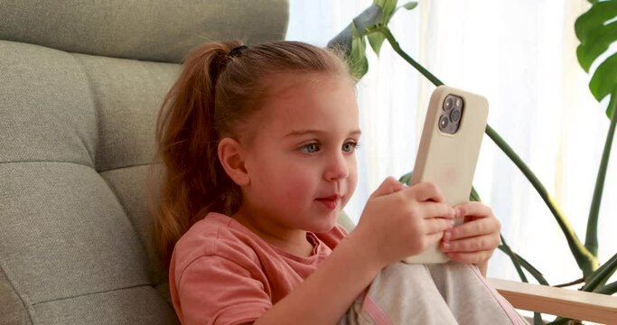 Cute baby girl sits chear and looks on smartphone. Child is talking via video link