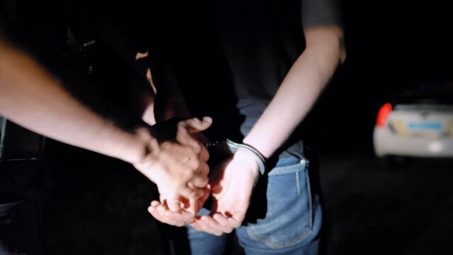 Police officer puts handcuffs on arrested man at night and leads him to a police car