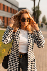 Beautiful young glamour blonde girl with style sunglasses in fashion clothes with a handbag walks in the city at sunset