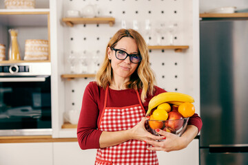 A housewife holding bowl with fresh fruits in kitchen at home.