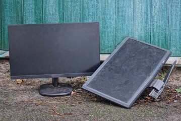 two old black computer monitors standing on the street on gray asphalt against a green wall