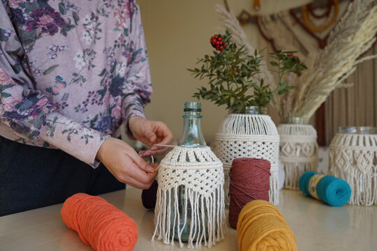 A woman decorates a bottle glass candlestick in the macrame style weaving,fir branches and rowan berries for home.Handmade decor in eco-style,hands close-up. Christmas, New Year, New hobby concept.