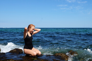 smiling red-haired teen girl in a black swimsuit sits on the seashore against the background of the...