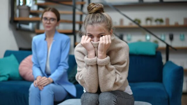 Portrait of introverted teenage girl looking at camera covering lower face with sweater sitting at psychologist appointment. Sad Caucasian therapist shaking head at background. Adolescence concept