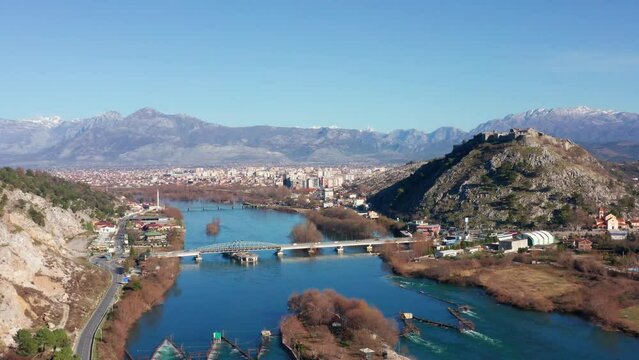 Buna (Bojana) river near Shkoder (Shkodra, Skadar), under Rozafa hill in Albania. Bridge across blue stream, with an islet (ait, eyot) in the foreground, under clear sky, on a sunny day.