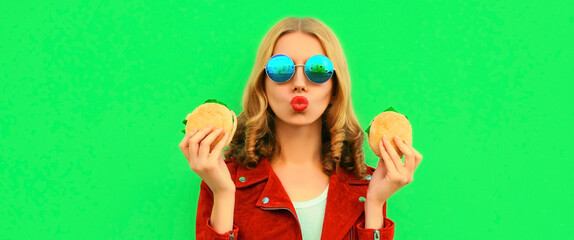 Portrait of stylish young woman eating tasty big burger fast food on green colorful background