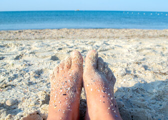 Two bare feet of caucasian woman soiled in sand with shell rock on sandy sea shore on sunny summer...