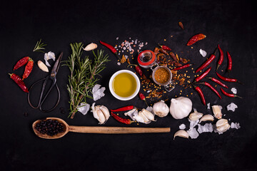 sprigs of rosemary, a metal scissor, a bowl with olive oil, juniper berries and various spices in the foreground, on a textured black background. View from above