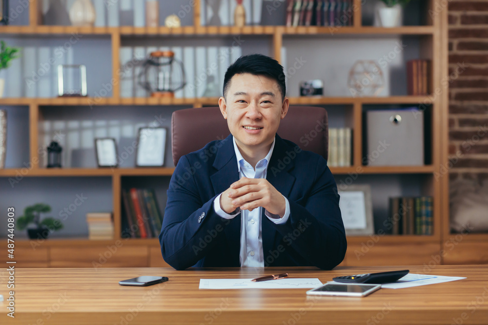 Wall mural successful asian broker businessman looks at camera and smiles, man at work sitting at table in clas