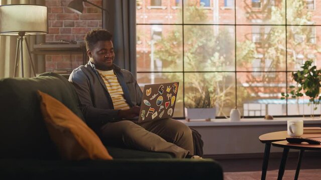 Young Handsome Black Man Working from Home on Laptop Computer in Sunny Stylish Loft Apartment. Creative Male Checking Social Media and Browsing Internet on a Sofa. Urban City View from Big Window.