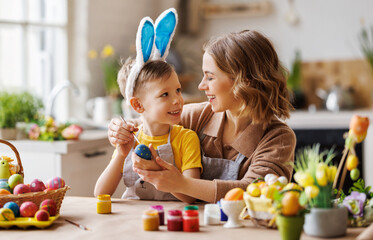 Loving young mother teaching happy little kid son to decorate Easter eggs while sitting in kitchen