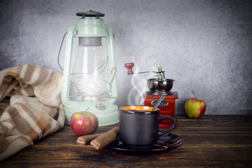 Kerosene lamp and black cup of coffee with saucer and spoon, with steam, coffee grinder, chocolate bars and apples on wooden table on gray background, brown checkered blanket nearby
