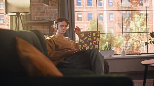 Young Handsome Man Sitting on Sofa and Using Laptop Computer in Sunny Stylish Loft Apartment. Creative Designer Wearing Cozy Yellow Sweater and Headphones. Urban City View from Big Window.