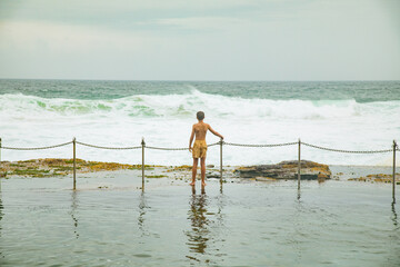 Boy standing at the fence on the edge of the Bogey Hole in Newcastle, NSW on an overcast day with...