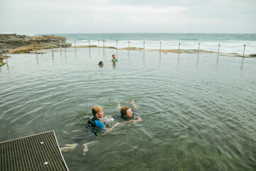 Fototapeta premium Brothers swimming at the Bogey Hole in Newcastle, NSW on an overcast day with rough surf