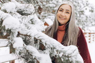 Senior woman with gray hair in a knitted sweater and scarf, sincerely smiling holding a Christmas...