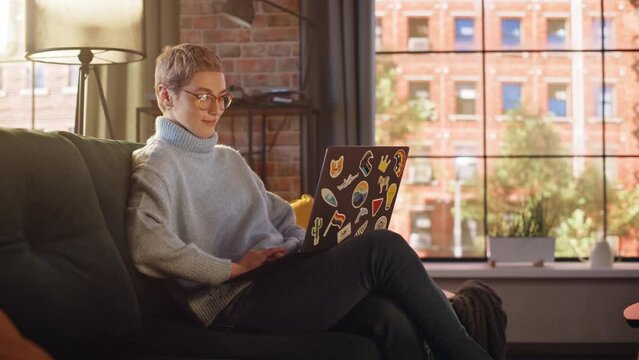 Young Beautiful Woman Working from Home on Laptop Computer with Stickers in Sunny Stylish Loft Apartment. Creative Female Checking Social Media, Browsing Internet. Urban City View from Big Window.