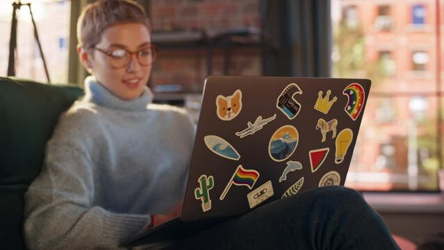 Stylish Female Using Laptop Computer with Diverse LGBT and Lifestyle Stickers on the Back. Young Creative Woman Sitting on a Couch, Typing, Browsing Internet and Checking Social Media at Home.