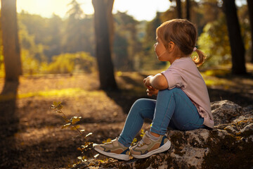 Young little girl sits on a rock in the forest and looks into distance