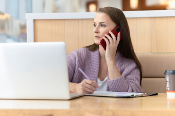 serious mature adult middle age woman freelancer working in workplace at computer, laptop, communicating on the Internet, using smartphone, mobile, signing documents. remote work. modern office