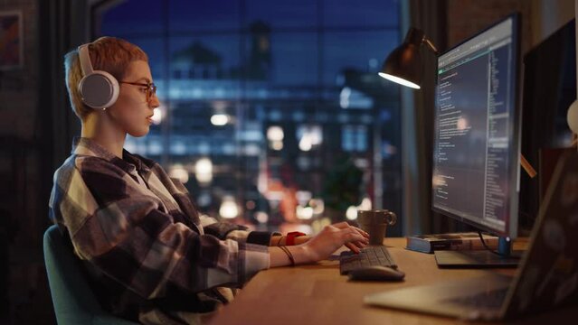 Young Woman Writing Code on Desktop Computer in Stylish Loft Apartment in the Evening. Creative Female Wearing Headphones, Working from Home on Software Development. Urban City View from Big Window.