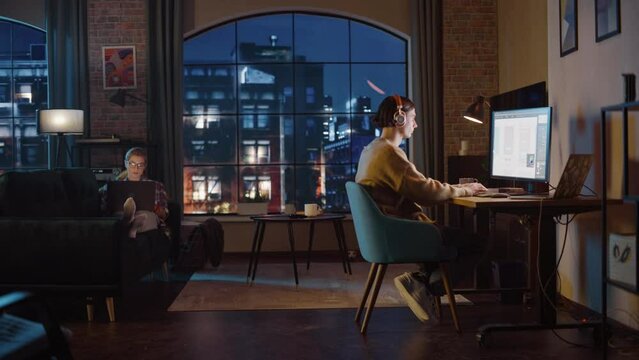 Young Couple Spending Time at Home, Working on Computers from Their Stylish Loft Apartment in the Evening. Female Resting on the Sofa and Browsing Social Media on Laptop. Man Designing UX Interface.
