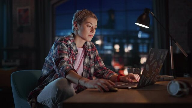 Young Attractive Woman Using Laptop Computer in Stylish Loft Apartment in the Evening. Creative Female Smiling, Checking Social Media, Browsing Internet. Urban City View from Big Window.