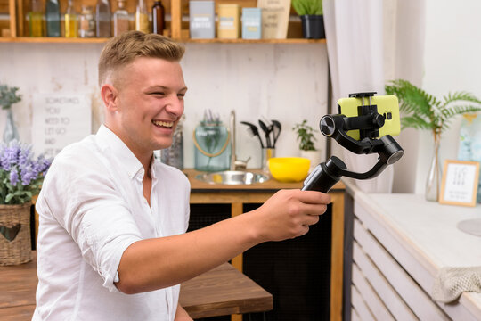 Blond European laughing guy filming a video blog on a mobile device using an electronic image stabilizer in the kitchen. A blogger shoots stories for Instagram. Blogger making a video for tiktok
