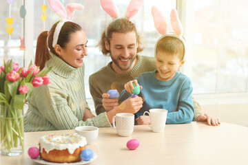 Happy parents with their little son in bunny ears cracking Easter eggs at table