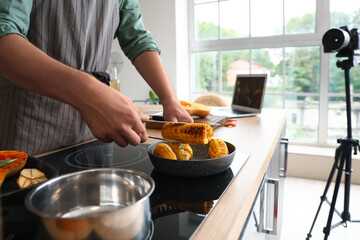 Young man cooking corn and recording video tutorial in kitchen