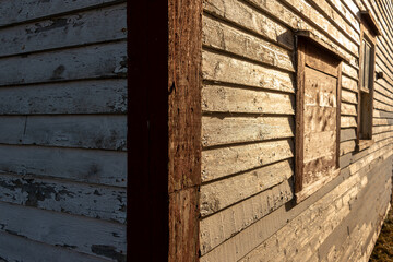 The exterior corner of an old red and white color wooden shed with multiple windows. The paint is peeling on the clapboard siding. One window is single hung glass and the other has a wooden shutter. 