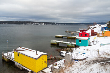 Fototapeta na wymiar Colorful and bright painted wooden fishing sheds or boathouses near the ocean with white snow covering the ground. The buildings are red, blue, and yellow in color. The sky is dark grey and cloudy. 