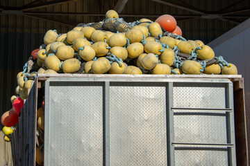 A commercial fishing net piled in a metal storage container. The industrial net is a green braided rope with plastic cork floats or small yellow and orange color buoys. The nets have edge markers.