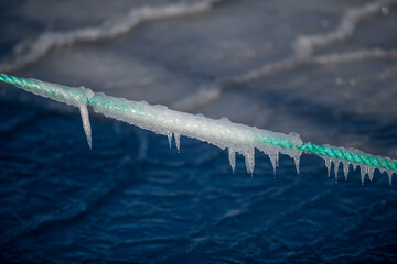 A braided green fishing rope hangs over a water surface with a skim of ice on top. There's thick...