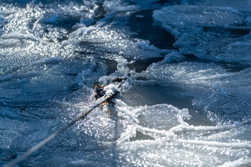 Naklejka premium A braided green fishing rope hangs over a water surface with a skim of ice on top. There's thick ice on the top section with snow and frosty ice crystals hanging from the green polypropylene line. 