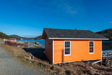 Fototapeta na wymiar The exterior wall of an orange color wooden boathouse. The building is made of wood cape cod siding. There are two closed glass windows with a black roof. The background is blue sky and ocean. 