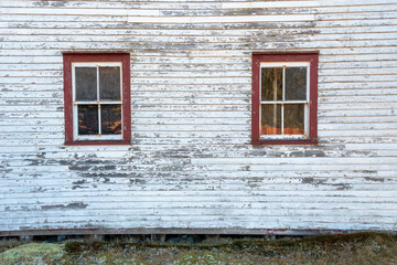 The exterior wall of a vintage white shed with two single hung closed windows. The trim on the windows is a vibrant red. The white paint is peeling on the wood siding of the storage building. 