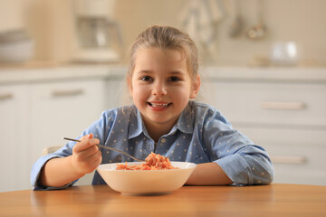 Cute little girl eating tasty pasta at table in kitchen