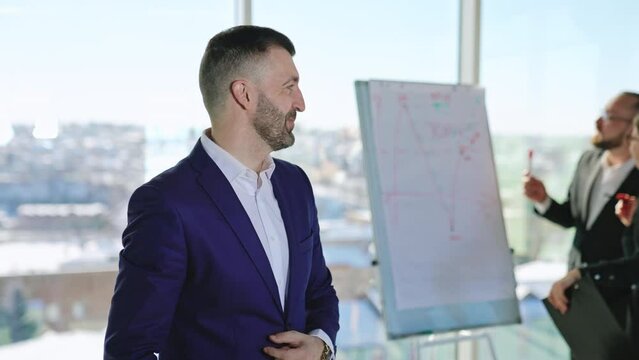 Mid-aged bearded man in suit talks firstly to camera and then aside. Portrait of a confident male business person crossing hands on his chest. Office employees at backdrop in blur.