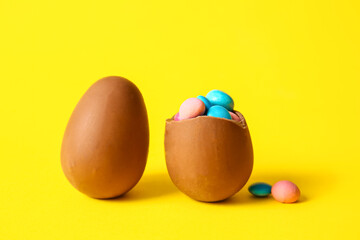 Broken chocolate Easter egg with different candies on yellow background, closeup