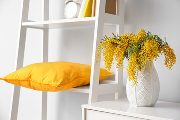 Vase with mimosa flowers on chest of drawers and shelving unit near light wall