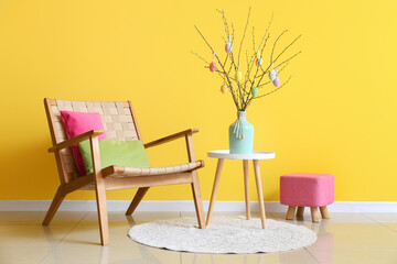 Tree branches with Easter eggs in vase on table, armchair and pouf near yellow wall