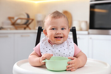Cute little baby with bowl in high chair at kitchen