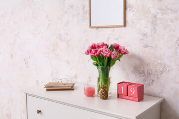 Vase with tulips, books, eyeglasses and cube calendar on chest of drawers near light wall