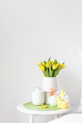 Vase with tulips, cups and Easter rabbits on table near light wall