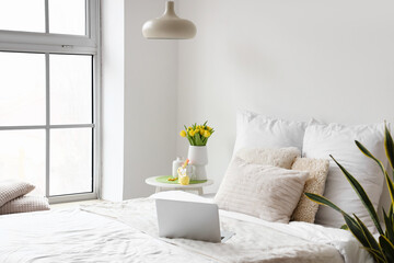 Comfortable bed with modern laptop, vase with tulips and Easter decor on table near light wall