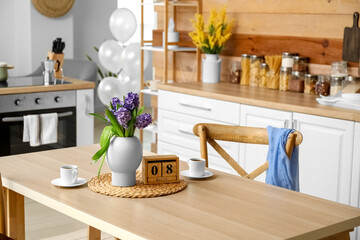 Cube calendar with date 8 MARCH, flowers and cup of coffee on dining table in kitchen. International Women's Day celebration