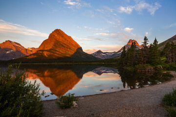 Beautiful dawn in mountains. Orange rocks reflect in calm water of the Swiftcurrent lake in the Glacier National Park, Montana