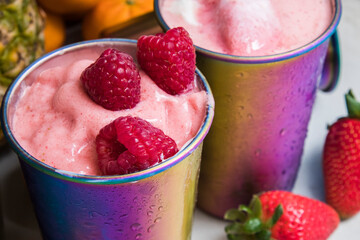 Metal rainbow colored cups with fresh fruit smoothies on kitchen counter with oranges, strawberries, limes, and pineapple