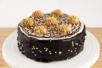 A whole chocolate cake covered with icing, decorated with cream, stands on a white stand on a wooden table.